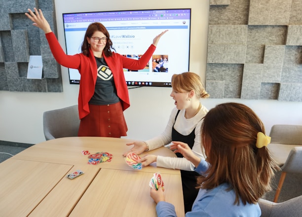 Four people are engaged in a lively game around a wooden table. One person stands with raised arms, while three others are seated, focusing on colorful cards in their hands. There is a computer monitor in the background displaying a webpage, and the room features a modern design with gray acoustic panels on the walls.